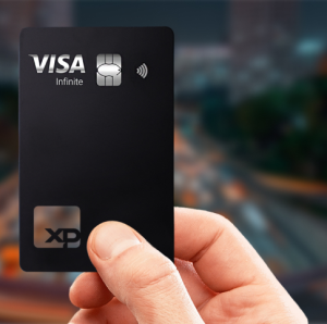Mão segurando cartão preto premium VISA Infinite XP com chip metálico e símbolo de pagamento por aproximação, em frente a um fundo urbano desfocado.
